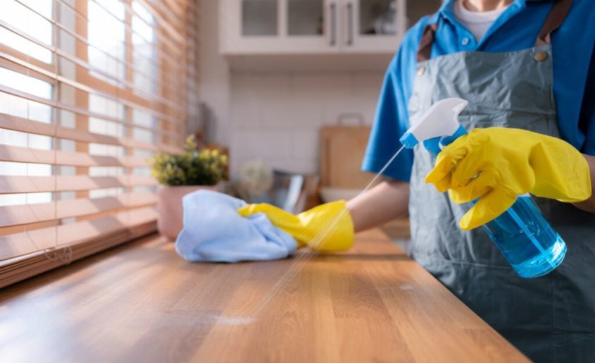 Cleaning service. Close-up of woman in apron and rubber gloves cleaning wooden table with spray